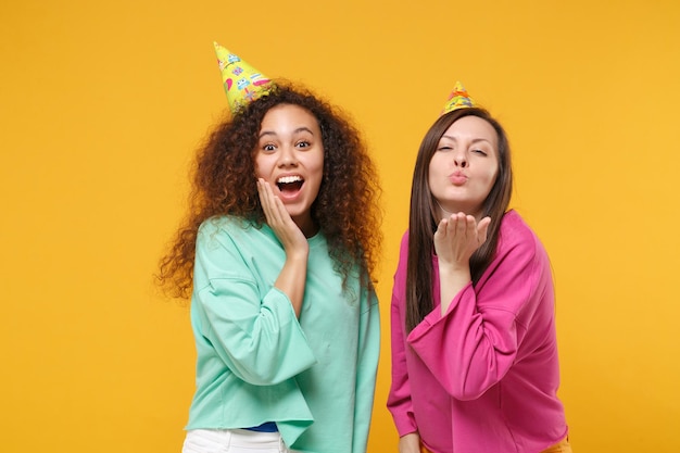 Due belle donne amiche ragazze afroamericane europee in abiti rosa verdi, cappelli di compleanno in posa isolati su sfondo giallo. Concetto di stile di vita delle persone. Mock up spazio di copia. Soffiando invia un bacio d'aria.