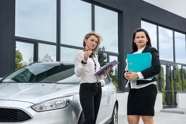 Due belle donne che si stringono la mano vicino alla nuova auto in piedi fuori e sorridono