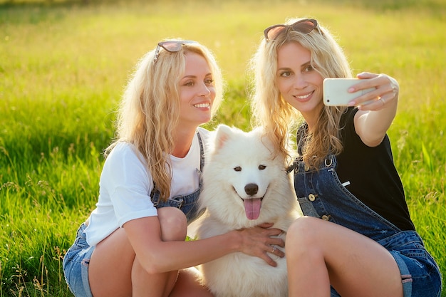 Due belle e affascinanti donne gemelle bionde con un cane samoiedo lanuginoso bianco seduto e facendo selfie al telefono nel parco.