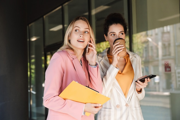 Due belle giovani donne d'affari sorridenti che camminano all'aperto per le strade della città, parlano mentre bevono caffè e usano i telefoni cellulari