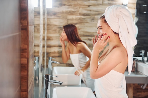 Due belle giovani donne in piedi in bagno vicino allo specchio insieme e pulendo i loro volti.
