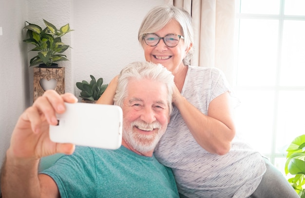 Due belle persone anziane si rilassano abbracciate sulla poltrona colorata guardando il cellulare per un selfie. Momenti positivi. Uomo con barba e capelli bianchi.