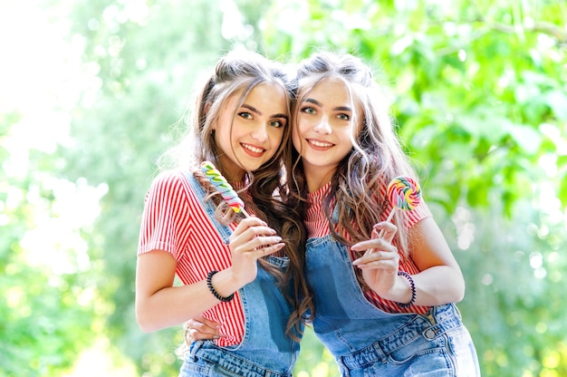 Due belle ragazze gemelle che abbracciano e ridono in tuta di jeans con lecca-lecca