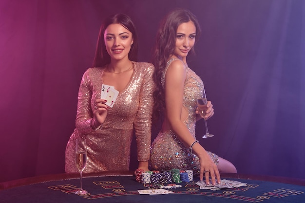 Due bellissime donne che mostrano le carte mentre posano al tavolo da gioco nel casinò Sfondo nero fumo con retroilluminazione colorata Poker d'azzardo Primo piano