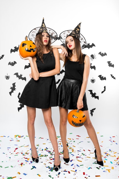 Due bellissime ragazze che indossano abiti neri, cappelli da strega e tacchi alti tengono le zucche di halloween sullo sfondo con i pipistrelli.