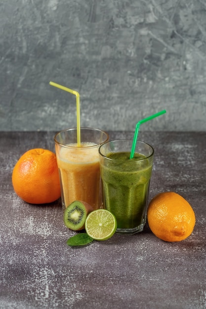 Due bicchieri alti di succo d'arancia e un frullato banana-arancia kiwi e spinaci circondati da metà di frutta su uno sfondo grigio cemento. Il concetto di perdere peso e una corretta alimentazione.