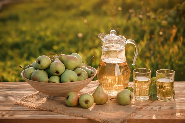 Due bicchieri con succo di pera e cesto con pere su tavola di legno con sfondo naturale del frutteto alla luce del tramonto Composizione di frutta vegetariana