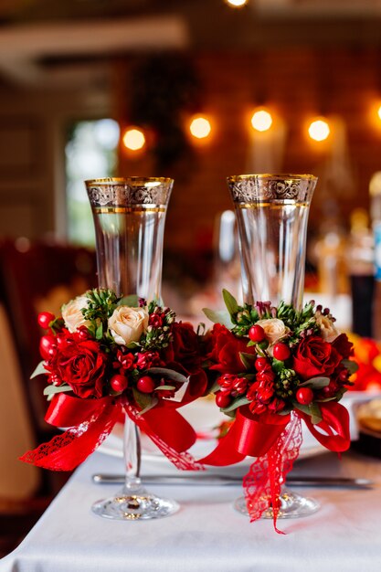 Due bicchieri da sposa vuoti, decorati con verde, rose rosse e nastro, in piedi sul tavolo del banchetto