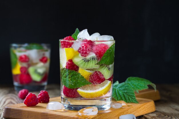 Due bicchieri di acqua minerale fresca, con bacche e frutti (lampone, limone, kiwi, ciliegia) e ghiaccio su sfondo nero