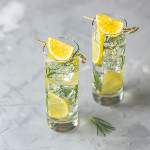 Due bicchieri di bevanda rinfrescante al limone e lime con cubetti di ghiaccio in calici di vetro su uno sfondo grigio chiaro Cocktail estivo di soda al limone fresco con fuoco selettivo al rosmarino