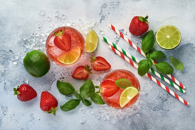 Due bicchieri di punch e ingredienti freschi per fare limonata, acqua detox infusa o cocktail. Fragole, lime, menta, basilico, cubetti di ghiaccio e shaker su pietra grigia o fondo in cemento. Vista dall'alto.