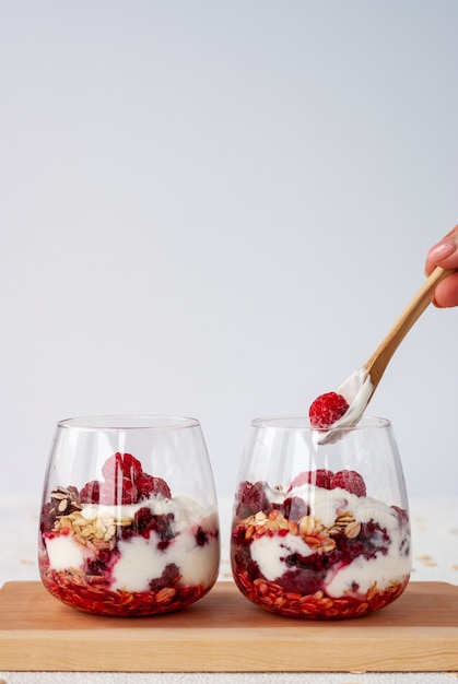 Due bicchieri di yogurt, fiocchi d'avena e lamponi con un cucchiaio di legno in piedi insieme su una tavola di legno