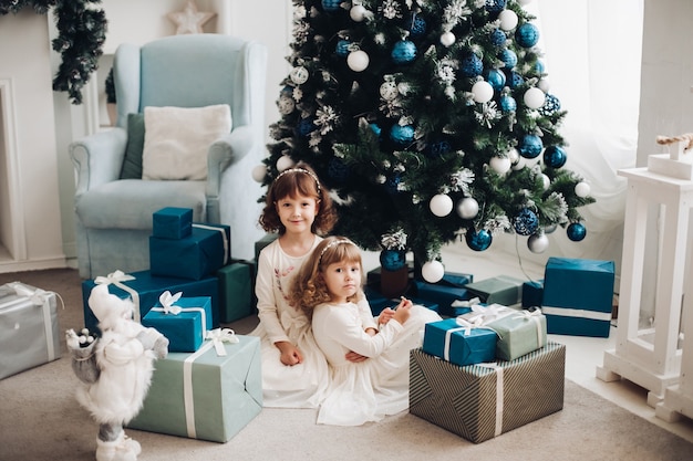 Due bionde bambine in abiti bianchi seduti vicino a pile di regali sotto l'albero di Natale