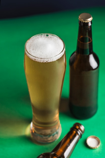 Due bottiglie di birra scure, un bicchiere di birra e schiuma su uno sfondo verde e blu.