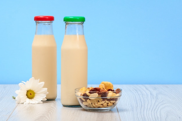 Due bottiglie di latte fresco con tappo rosso e verde, ciotola di vetro con muesli e fiori di camomilla su sfondo blu