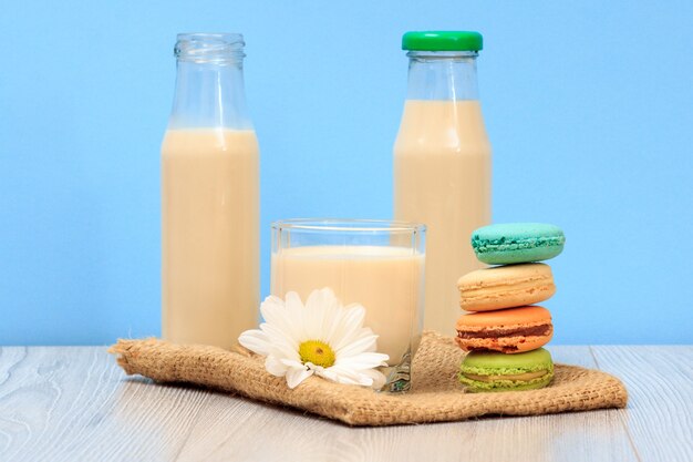 Due bottiglie e un bicchiere di latte fresco con fiori di camomilla e amaretti su tela di sacco e sfondo blu.
