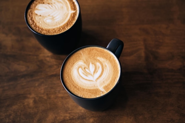 Due caffè cappuccino con latte art su tavola di legno Tazze nere e bella schiuma di latte Soft focus