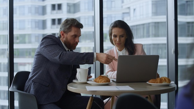 Due colleghi alla ricerca di computer portatile che lavorano online nella caffetteria dell'ufficio di vetro Uomini d'affari fiduciosi che prendono appunti spiegando la strategia finanziaria Colleghi che lavorano insieme Concetto di freelance