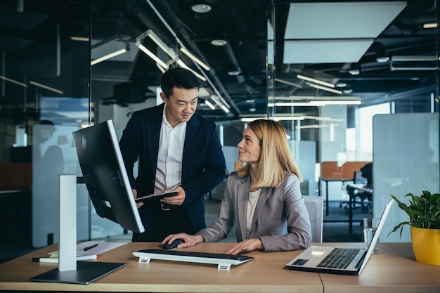 Due colleghi asiatici maschi e femmine in un ufficio moderno una donna mostra il lavoro svolto sul monitor consulta e discute