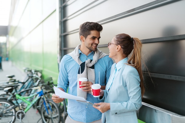 Due colleghi sorridenti hanno vestito la condizione elegante all'aperto e la tenuta dei documenti mentre bevono il caffè.