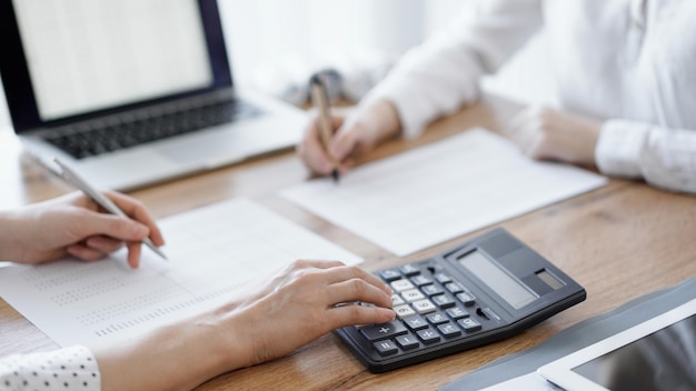 Due contabili che usano un computer portatile e una calcolatrice mentre contano le tasse alla scrivania di legno in ufficio. Lavoro di squadra nell'audit aziendale e nelle finanze.
