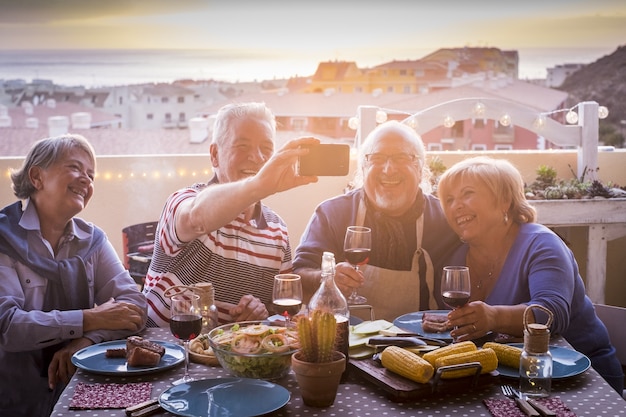 Due coppie anziane allegre che cenano alla festa in terrazza e si fanno selfie utilizzando lo smartphone