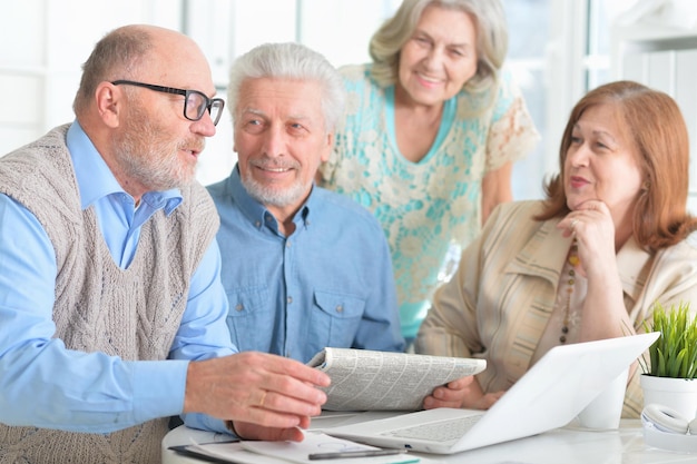 Due coppie senior che si siedono al tavolo che lavorano con il computer portatile e che leggono il giornale