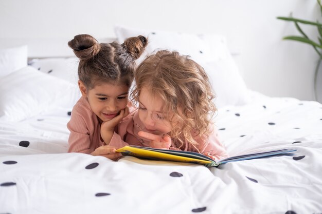 Due diversi bambini ragazza sorelle amici in pigiama libro di lettura sdraiato su biancheria da letto bianca sul letto di casa.