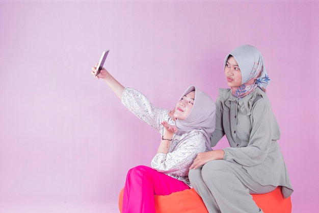 Due donne adolescenti in hijab che prendono selfie con i telefoni cellulari seduti su sedie di fagioli arancioni isolate p