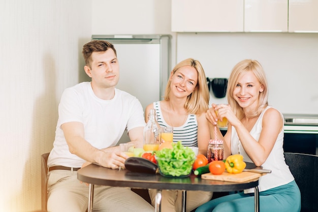 Due donne adorabili felici e un bell'uomo seduto in cucina, sorridente guardando la fotocamera. Gli amici cucineranno la cena dalle verdure. Stile di vita, dieta e concetto di cibo sano.