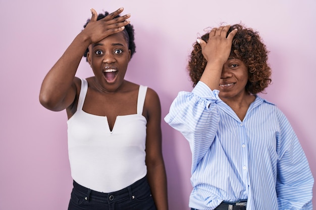 Due donne africane in piedi su sfondo rosa sorprese con la mano sulla testa per errore, ricordano l'errore. dimenticato, cattivo concetto di memoria.