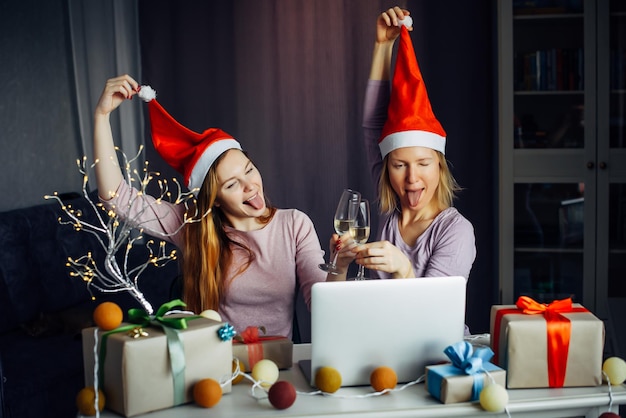 Due donne allegre con cappelli di Babbo Natale che bevono champagne e si congratulano a distanza con gli amici per Natale seduti a tavola con il laptop. Le amiche si divertono a capodanno all'interno di casa.