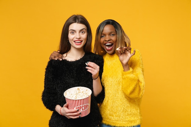 Due donne amiche europee e afroamericane in abiti gialli neri tengono un secchio di popcorn isolato su uno sfondo arancione brillante, ritratto in studio. Concetto di stile di vita delle persone. Mock up spazio di copia.