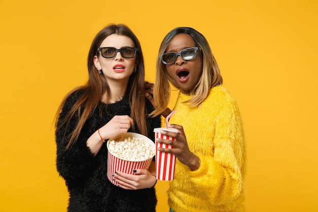 Due donne amiche europee e afroamericane in abiti gialli neri tengono un secchio di popcorn isolato su uno sfondo arancione brillante, ritratto in studio. Concetto di stile di vita delle persone. Mock up spazio di copia.