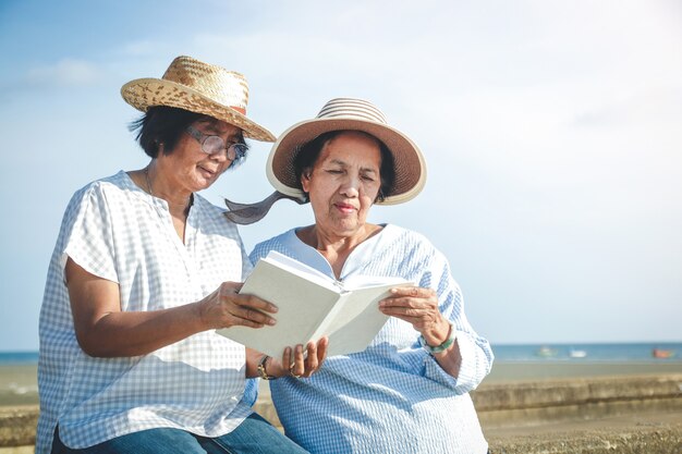 Due donne anziane asiatiche seduti in spiaggia a leggere un libro