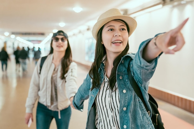 due donne asiatiche amici turisti che cercano posizioni guardando il segno nel passaggio eccitato che punta. ragazze che trovano l'uscita nel corridoio della stazione sindacale. curiosa femmina gioiosa che mostra il modo della sorella.