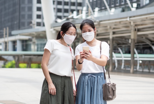 Due donne asiatiche che indossano una maschera medica ogni volta fuori casa, come nuova tendenza normale e autoprotezione, usando lo smartphone per selfie o cercando informazioni commerciali