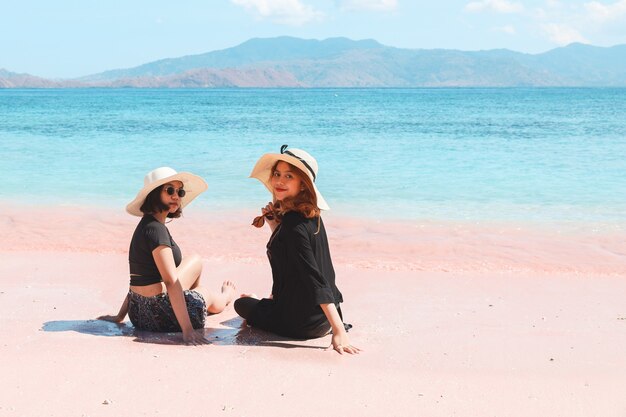 Due donne asiatiche in cappello estivo seduto e guardando la telecamera in spiaggia di sabbia rosa a Labuan Bajo