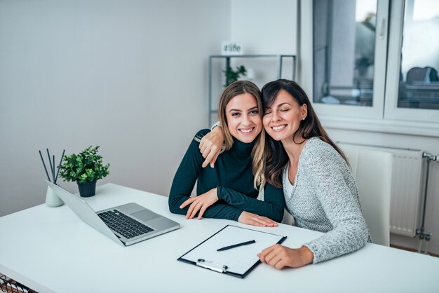 Due donne casuali di affari che abbracciano mentre si siedono sul lavoro. Amici che lavorano insieme.