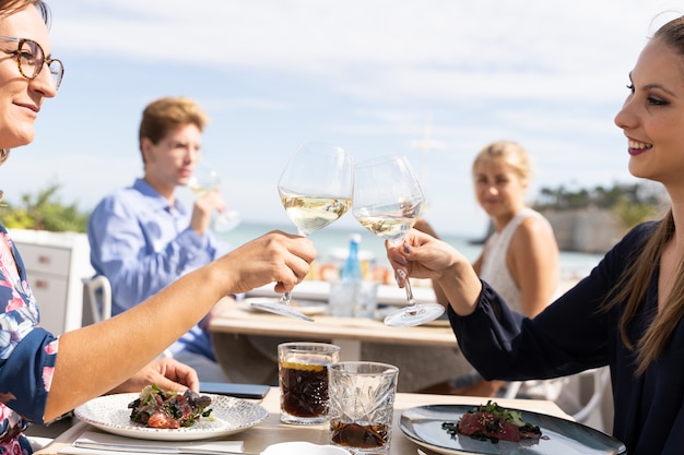 Due donne che tostano con bicchieri di vino bianco seduti a un tavolo in un ristorante sulla spiaggia