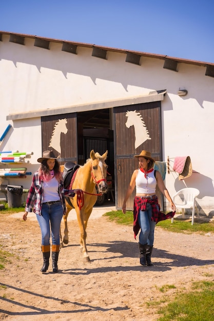 Due donne cowgirl che escono sorridenti tirando fuori un cavallo da una stalla, con abiti sudamericani, stile di vita di due che lavorano in una stalla