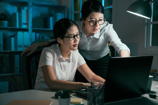 due donne d'affari asiatiche che lavorano discutono con il computer portatile in ufficio serale. dipendenti di sesso femminile che guardano lo schermo del notebook puntano il dito sul monitor a parlare. signora manager che insegna al tirocinante di notte