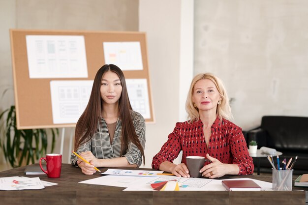 Due donne d'affari contemporanee fiduciose in abbigliamento casual intelligente che bevono alla scrivania mentre lavorano su un nuovo progetto di design in ufficio