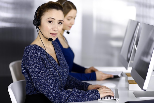 Due donne d'affari conversano con i clienti tramite cuffie, mentre sono sedute alla scrivania di un ufficio moderno. Diverse persone raggruppano in un call center. Telemarketing e servizio clienti.