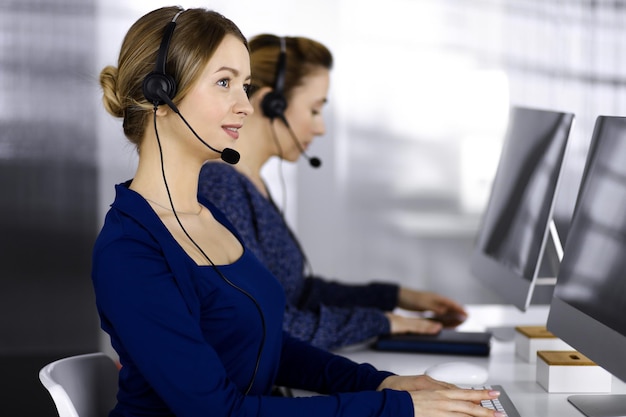 Due donne d'affari conversano con i clienti tramite cuffie, mentre sono sedute alla scrivania di un ufficio moderno. Diverse persone raggruppano in un call center. Telemarketing e servizio clienti.