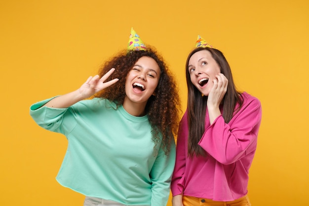 Due donne divertenti amiche ragazze afroamericane europee in abiti rosa verdi, cappelli di compleanno in posa isolati su sfondo giallo. Concetto di stile di vita delle persone. Mock up spazio di copia. Mostra il segno della vittoria.