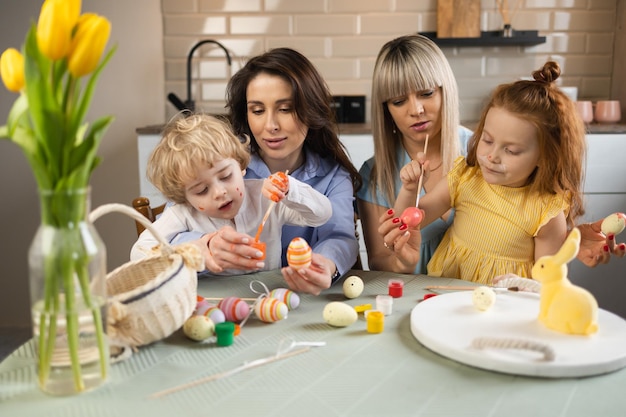 Due donne e i loro bambini sono seduti in cucina a tavola e dipingono le uova di Pasqua