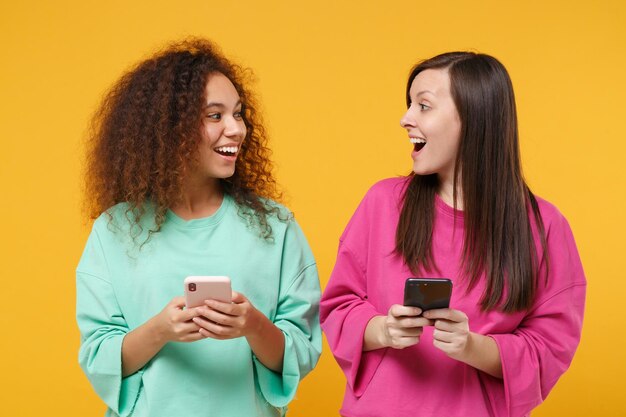 Due donne eccitate amiche ragazze afroamericane europee in abiti rosa verdi in posa isolati su sfondo giallo. Concetto di stile di vita delle persone. Mock up spazio di copia. Utilizzando il telefono cellulare, digitando il messaggio.