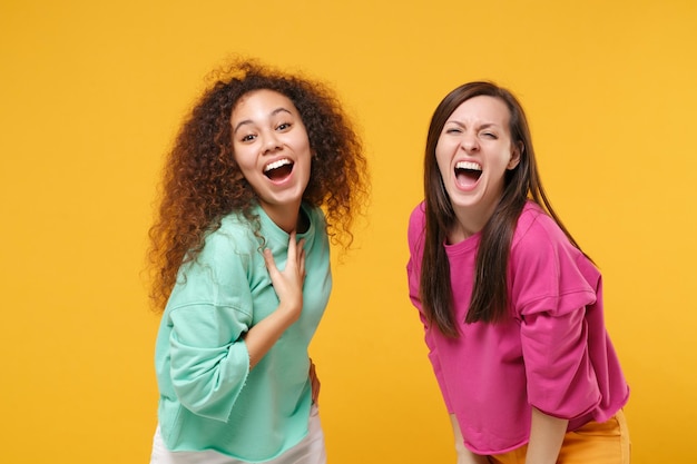 Due donne eccitate che ridono amiche ragazze europee e afroamericane in abiti rosa verdi in posa isolata su sfondo giallo arancione ritratto in studio. Concetto di stile di vita delle persone. Mock up spazio di copia.