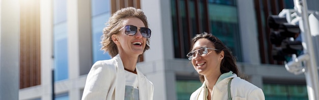 Due donne felici in abiti alla moda ridono e camminano fuori sullo sfondo di un edificio moderno